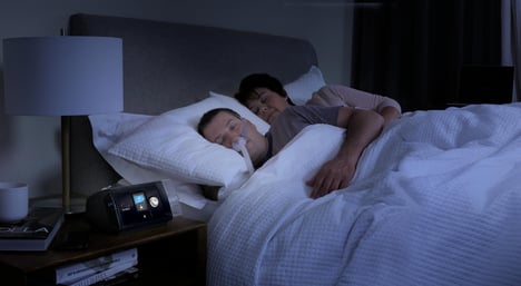 Man with cpap mask sleeping next to a woman, they are sleeping on their sides.